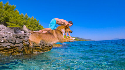 LOW ANGLE VIEW: Brown dog and young man simultaneously jump head first into refreshing crystal clear blue sea from a rocky Croatian seashore. They are enjoying summer holidays on sunny island of Hvar.