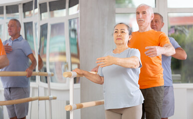 Wall Mural - Positive old lady with aged women and men engaged in group class perform exercise near ballet barre, standing in first position of ballet stance in modern dance studio