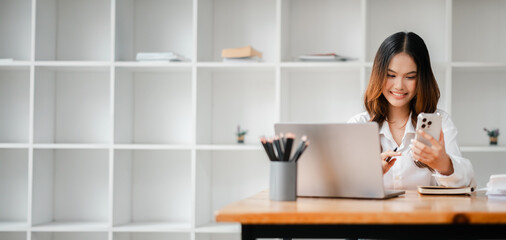 Wall Mural - A woman is sitting at a desk with a laptop and a cell phone. She is smiling and she is enjoying her work. The desk is cluttered with various items such as a cup, a book, and a potted plant