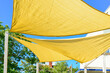 © Dolores  Harvey - Multiple large summer nylon patio sun shade umbrellas, sunny yellow color with wooden supports. The sun is shining through the umbrellas. The background is a bright blue sky, trees, and a building.