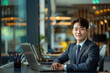 © NaphakStudio - A young Korean businessman in a suit, sitting at a desk with a laptop in a modern office, smiling confidently at the camera.