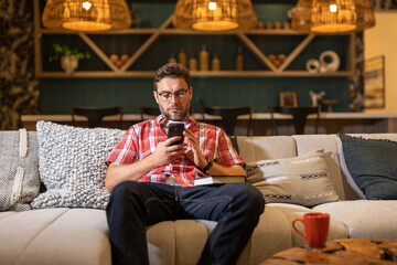 Handsome cheerful man indoors at home on sofa using mobile phone. Attractive caucasian man resting on sofa in apartment at night texting on call phone, surfing internet on smartphone.