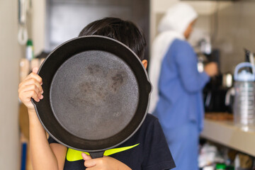 kids holding cast iron skillet with a background of his mom cooking in the kitchen, cocept of helping mom