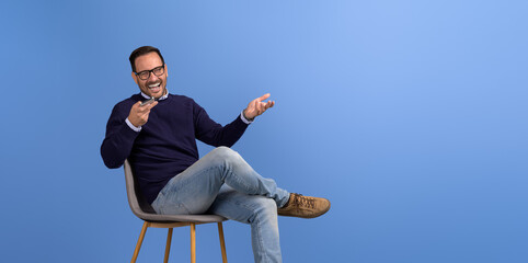 young businessman laughing and talking on speakerphone while sitting on chair over blue background