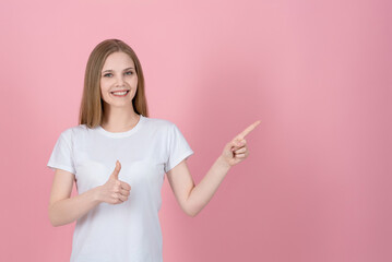 Wall Mural - Attractive caucasian young blonde woman in casual white t-shirt smiling, showing thumb up and pointing finger at empty copy space for advertising isolated on pink studio background