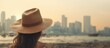 © Ilgun - A girl with a hat on her head gazes into the distance on the beach with buildings in the background one hand holding the hat and the other touching her neck. Copy space image