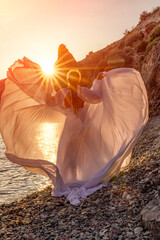 Wall Mural - Mysterious woman silhouette long hair walks on the beach ocean water, sea nymph wind listens to the wave. Throws up a long white dress, a divine sunset. Artistic photo from the back without a face