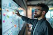 © Ilia Nesolenyi - A man wearing glasses is pointing to a whiteboard covered with flowcharts, graphs, and bullet points, along with sticky notes