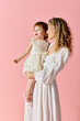 © LIGHTFIELD STUDIOS - A mother in a white dress lovingly holds her baby girl in a white dress on a soft pink background.