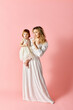 © LIGHTFIELD STUDIOS - A woman in white dress holds a baby girl in a white dress against a pink background.