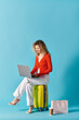 © LIGHTFIELD STUDIOS - Curly-haired woman sitting on suitcase, using laptop.