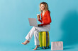 © LIGHTFIELD STUDIOS - Curly-haired woman sitting on suitcase, typing on laptop, and enjoying coffee.