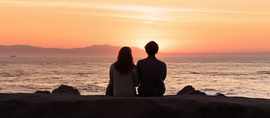 Wall Mural - Couple playfully sitting on a wall at sunset by the ocean seen from behind with copy space in the image