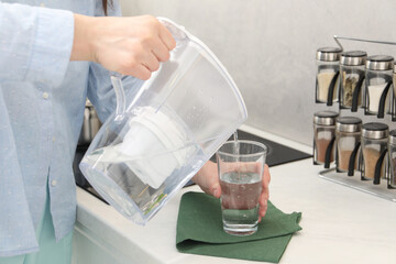 Wall Mural - Woman pouring water from filter jug into glass in kitchen, closeup