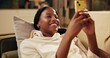 © peopleimages.com - Black woman, couch and laughing with smartphone for social media, online comic and funny chat with friends. Gen z girl, mobile and smile for internet joke with happy, meme and streaming video on sofa