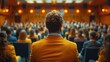 © Vilaysack - A presenter in a yellow jacket addresses a large audience in a conference hall, creating a focused and engaging atmosphere under warm lighting.