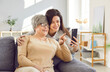 © Studio Romantic - Smiling happy adult child woman using mobile phone with cheerful mature senior mother parent sitting on sofa at home having online video call or watching news in social media. Family leisure concept.