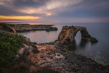 Wall Mural - Rocky coastline with a natural arch at punta Asparano, near Siracusa. Sunrise time, long exposure picture. June 2023