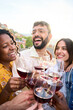 © CarlosBarquero - Vertical. Cheerful international friends toasting glasses of red wine together laughing at terrace bar outdoors. Close up cheers of smiling diverse people having fun at sunset party. Summer vibes