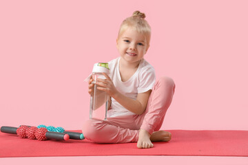 Poster - Cute little girl sitting on yoga mat with body rollers and bottle of water against pink background