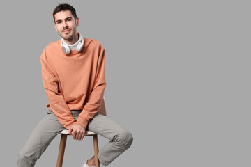 Wall Mural - Handsome young man in stylish peach sweater with headphones sitting on chair and posing against grey background