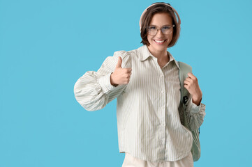 Wall Mural - Happy female student with backpack and headphones showing thumb-up on blue background