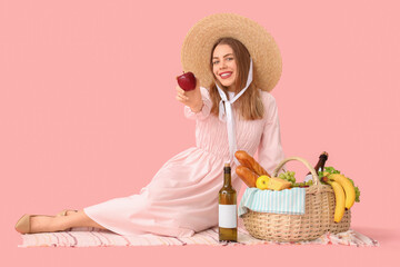 Poster - Beautiful young happy woman with food and bottles of wine for picnic in wicker basket sitting on pink background