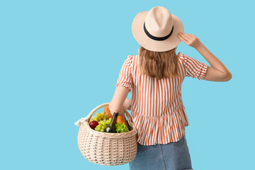 Wall Mural - Beautiful young woman with food and bottle of wine for picnic in wicker basket on blue background, back view