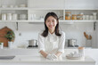 © Sourav Mittal - A young Japanese woman cleaning the kitchen counter in a white minimalist modern home interior, smiling and looking at the camera with copy space on the right side. In the style of a Japanese house