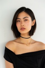 Wall Mural - young Asian woman with short, straight hair, wearing a black one-shoulder top and a gold choker necklace, white background