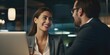 © vefimov - A woman and a man are sitting at a table, smiling at each other. The woman is wearing a suit and the man is wearing a suit and glasses. They are both looking at a laptop, which is open on the table