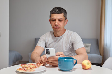 Sad upset Caucasian man wearing T-shirt using mobile phone watching videos in internet scrolling social networks expressing sadness and sorrow sitting at table in home interior