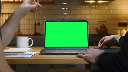 Two anonymous men working with laptop computer with green mock up screen on table in the kitchen pointing screen with hand. Co-workers working home office, remote work, technology concept 