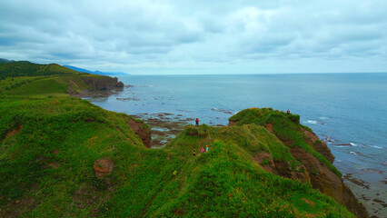 Wall Mural - Top view of tourists on rocky coast trail with green grass. Clip. Beautiful landscape of rocky coast with green grass and tourists on hike. Tourists ride on edge of rocky coast overlooking sea on