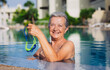 © luciano - Happy active senior woman doing sport in the outdoor swimming pool under the sun. Smiling woman holding her swimming goggles. Healthy lifestyle and retirement concept