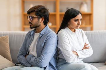 Indian young couple sits back-to-back on a couch, both with arms crossed, appearing to be in the midst of an argument in a bright living room.