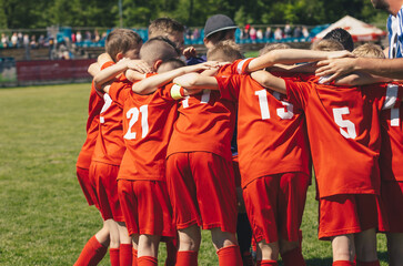 Wall Mural - Children's sports team with coach. Kids play sports match on stadium field. School boys in red jersey shirts huddling in a team. Parents sitting at the stadium and watching pupils in the background 