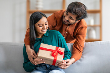 Wall Mural - Indian man sitting on a couch is handing a gift to a woman next to him. The woman appears surprised and delighted as she receives the gift