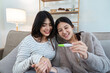 © itchaznong - Two happy gay lesbian lgbt women couple sitting on sofa celebrating positive pregnancy test. lgbtq concept
