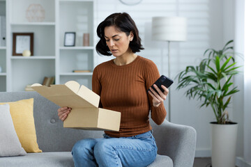 An upset young woman is sitting on the sofa at home, holding a mobile phone in her hand and worriedly looking into an open box of a parcel that she received by mistake