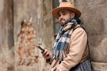 Wall Mural - Smiling man in warm scarf with smartphone near wall outdoors. Space for text