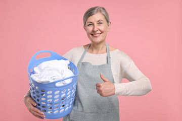 Canvas Print - Happy housewife with basket full of laundry showing thumbs up on pink background