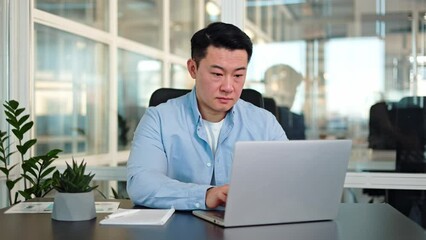 Focused asian man with neat hairstyle typing on keyboard of portable computer while working at office with glass walls. Busy representative answering emails and setting communication with partners.