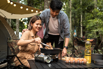 Happy Asian couple cooking food in a pan on tourist gas burner near the tent at camp. Camping and travel vacation concept.