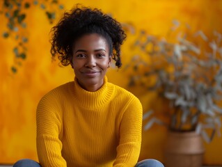 Wall Mural - A woman in a yellow sweater is smiling and sitting in front of a yellow wall. Scene is cheerful and warm