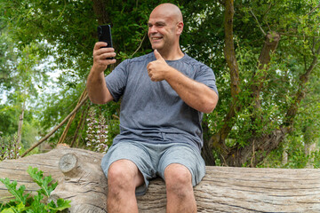 Wall Mural - A smiling man sitting on the trunk of a tree, using the phone