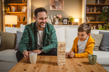 Father with cup of hot drink and son play board game together at home