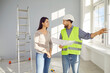 © Studio Romantic - Builder or construction foreman with paper plan talking to homeowner. Friendly, smiling man in hardhat and uniform vest talking to young woman while standing in room with ladder in new house