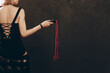 © vladdeep - Slender Woman holding Black and Red Leather Flogging Whip in hand in dark interior, view from back