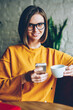 © BullRun - Half length portrait of positive beautiful businesswoman in spectacles smiling at camera sending notification with good news via high speed internet connection on smartphone resting in cafe interior